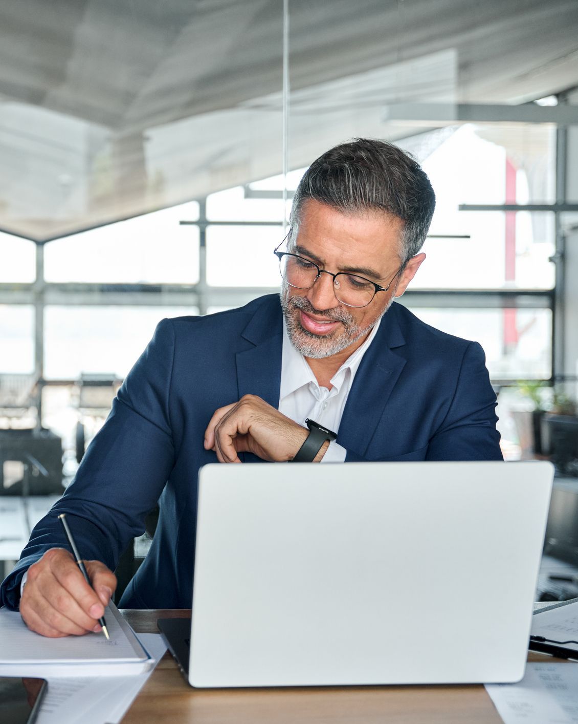 Businessman in suit writing in notebook and checking smartwatch at laptop