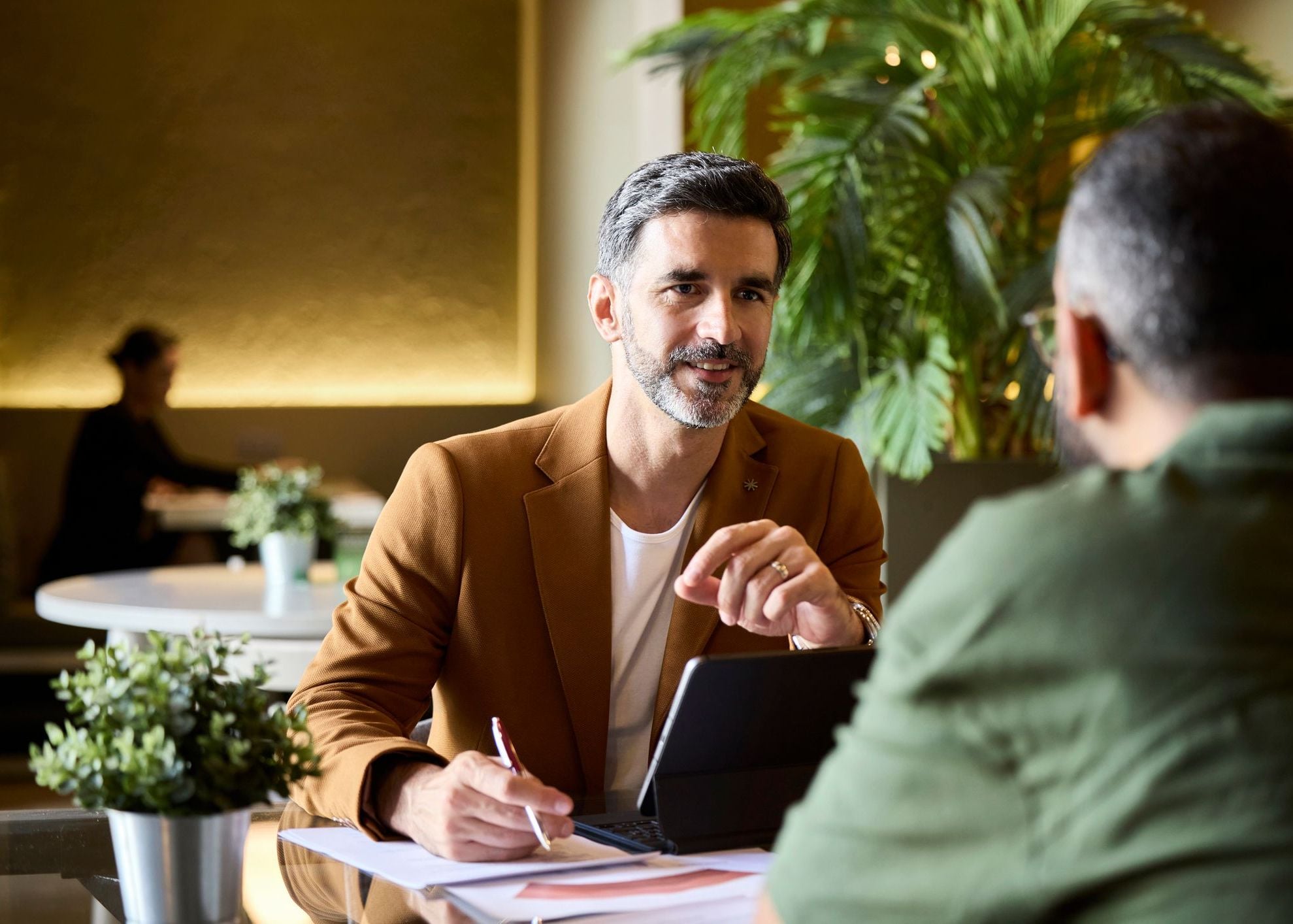Business meeting with two men at a table, one in a brown suit, in a modern office with plants.